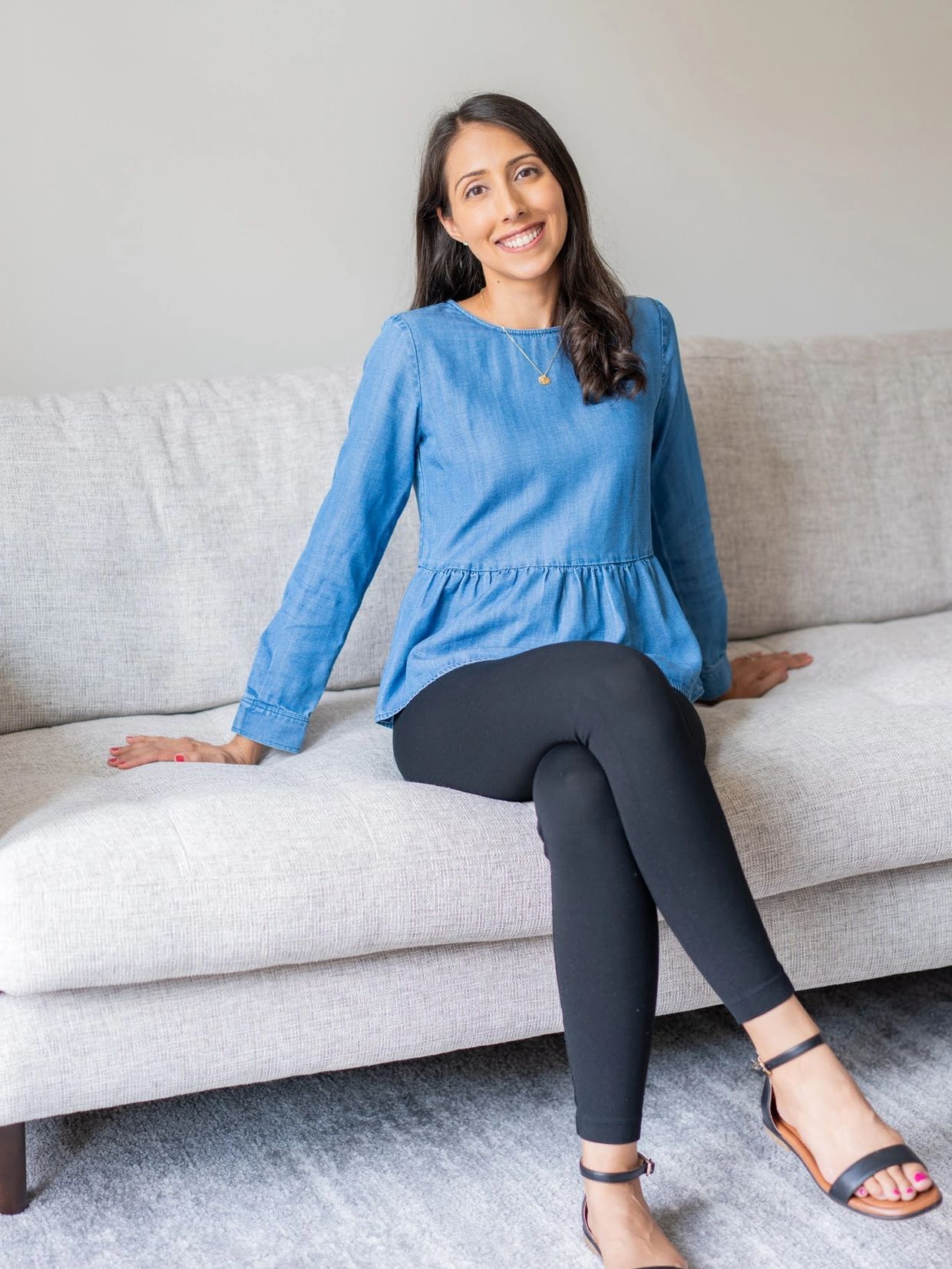 Smiling woman in blue top sitting on a beige sofa in a bright room.