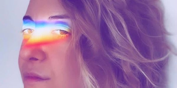 A queer woman with light hair has an LGBTQIA+ rainbow light cast across her eyes and nose.