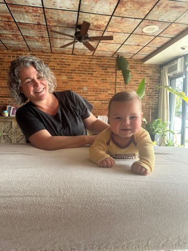 Smiling woman watches a happy baby lying on a bed in a cozy room.