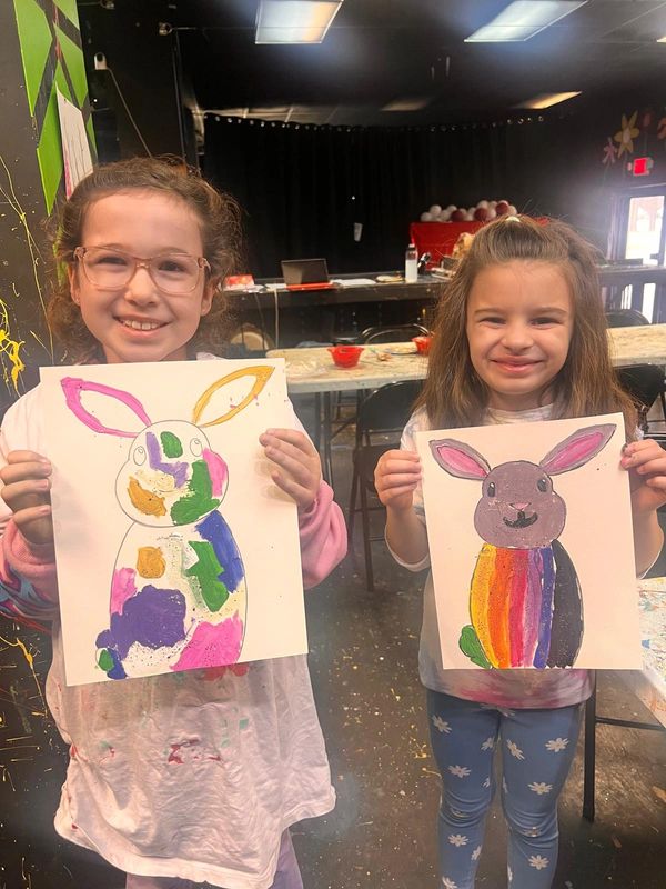 Two girls proudly display their colorful bunny paintings in an art studio.