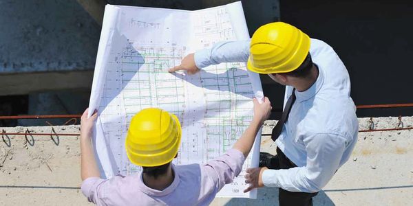 Two construction workers in yellow helmets reviewing blueprints at a building site.
