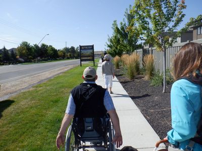 Photo from behind of a man in a wheelchair and a woman with a guide dog moving down a sidewalk.