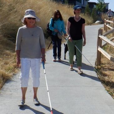 Photo of three women, two using white canes and one with a guide dog, walking down a trail.