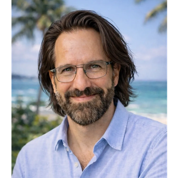 Smiling man with glasses and beard by the beach.