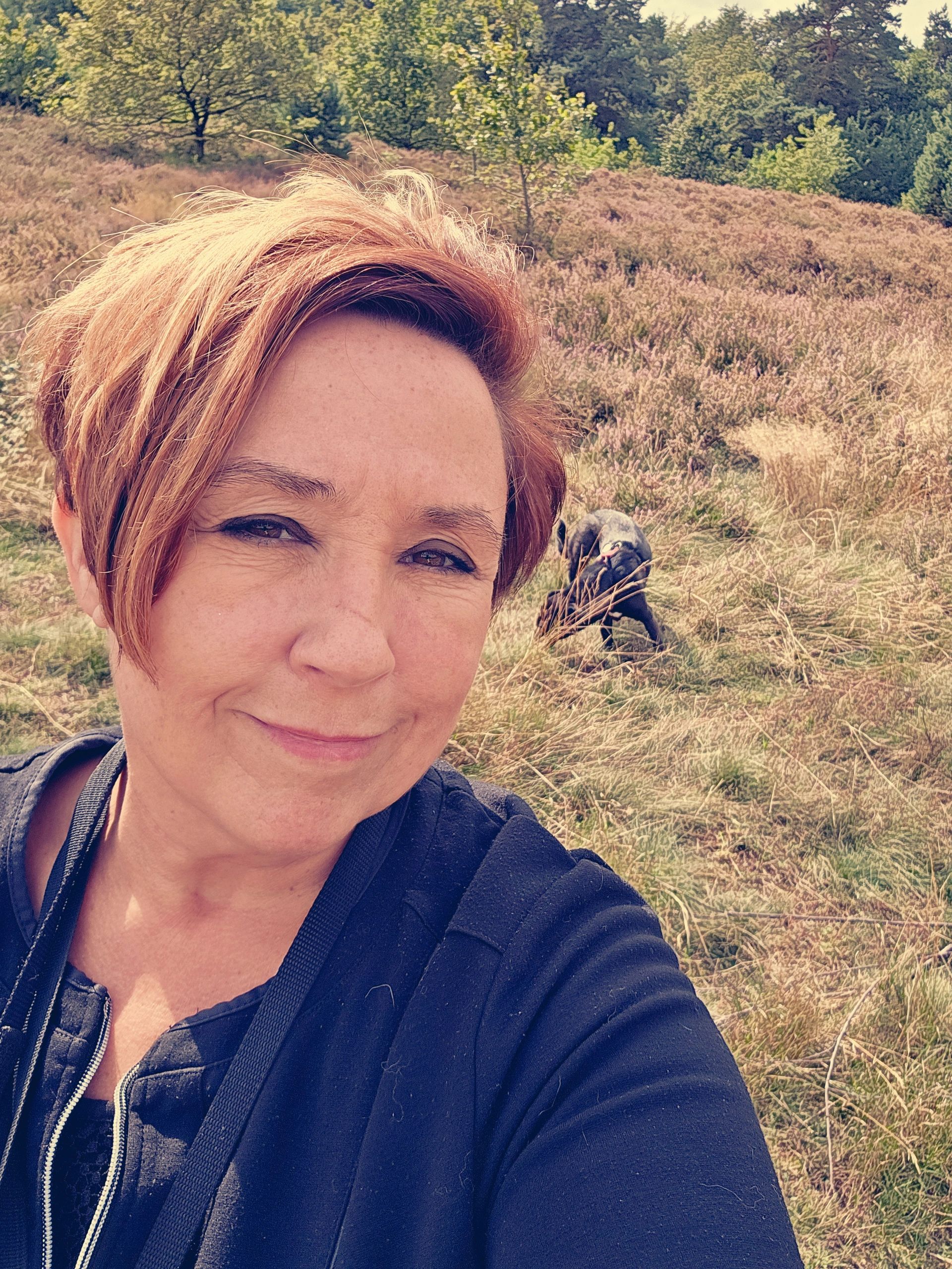 Woman taking a selfie outdoors with a dog in the background in a grassy field.