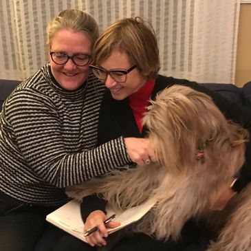 Two women with glasses petting a fluffy dog indoors.