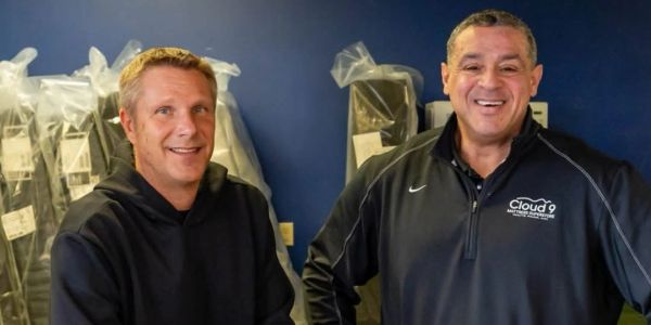 Two men smiling indoors with packaged items behind them.