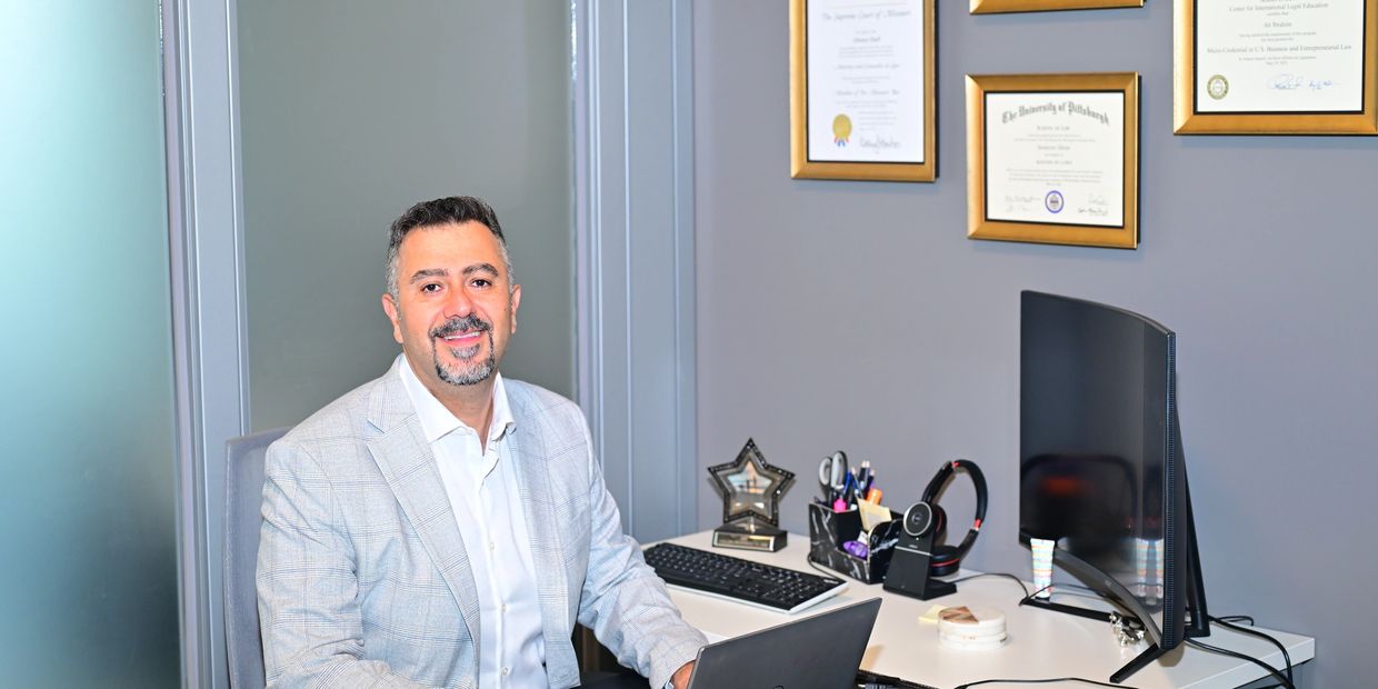 Man in light suit sitting at a desk with computer and certificates on wall.