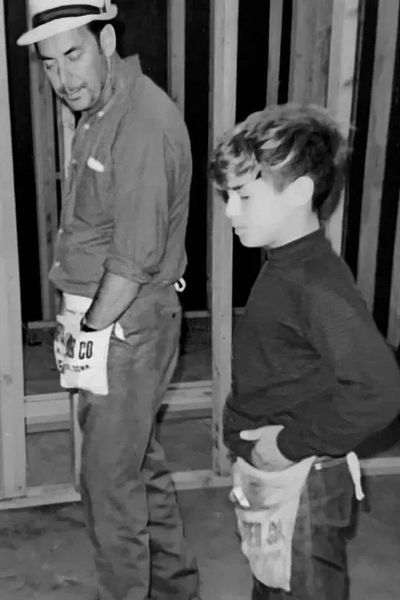 A man and boy working together on a construction site, wearing tool belts.