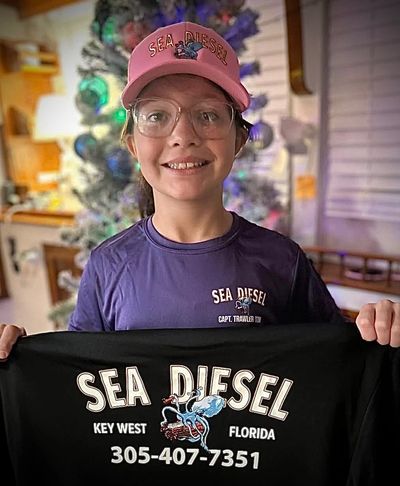 Smiling boy wearing Sea Diesel hat and shirt, holding a Sea Diesel sign indoors.