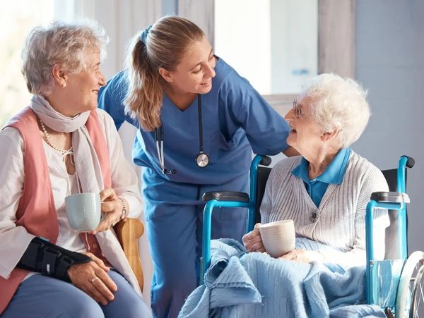Caregiver warmly interacts with two elderly women, one in a wheelchair, sharing a moment over tea.