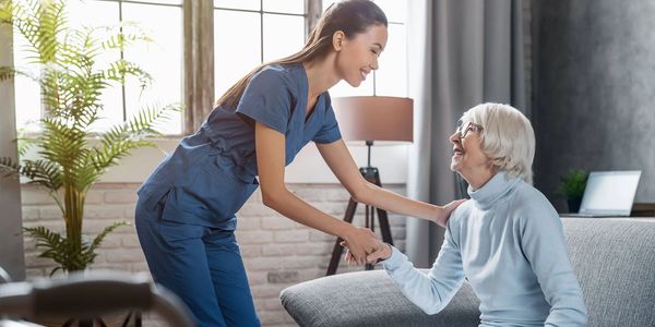 A nurse warmly assists a smiling elderly woman sitting on a couch.