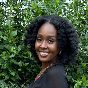 Smiling woman with curly hair posing outdoors in front of lush greenery.