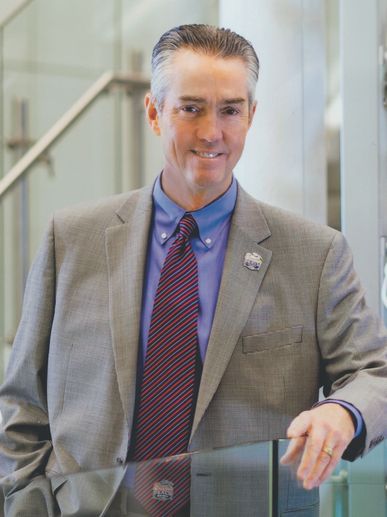 A middle-aged man in a suit with a striped tie smiles confidently indoors.