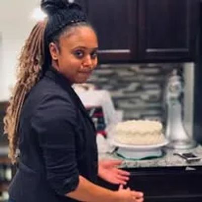 A woman with dreadlocks stands near a kitchen counter with a decorated cake.