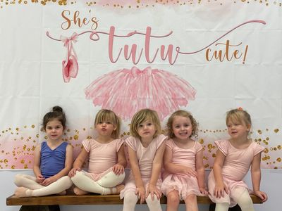 Five young ballerinas sitting on a bench in front of a tutu-themed backdrop.