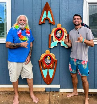Two men posing with colorful wooden wall art outside a blue building.