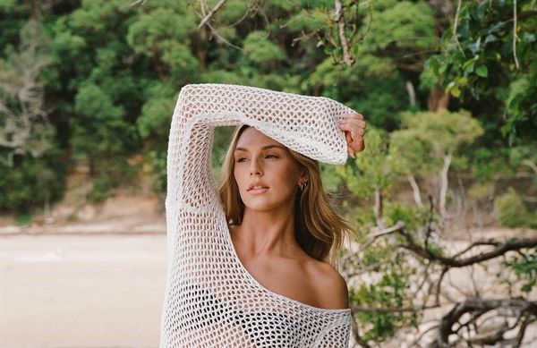 A woman poses outdoors wearing a white netted top, with a serene forest background.