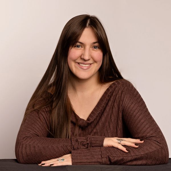 Young woman with long brown hair smiling in a brown sweater.
