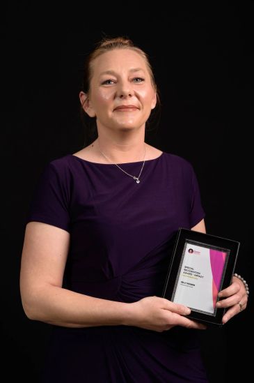 Woman in purple dress holding an award plaque against a black background.
