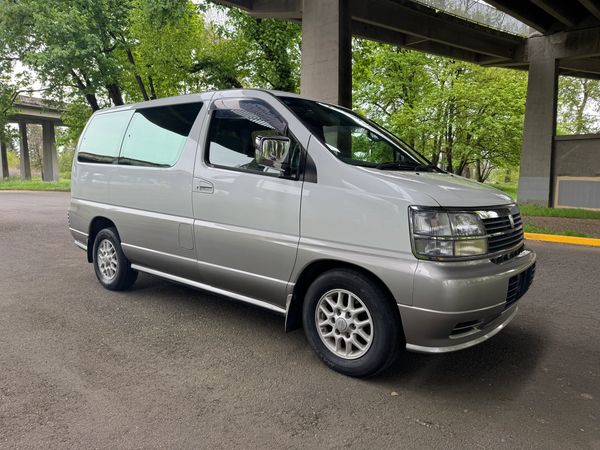 Silver and white passenger van parked under a bridge with trees in the background.