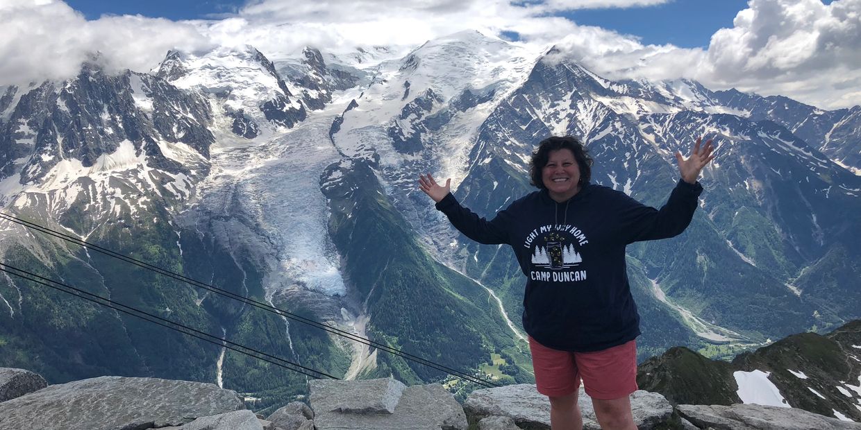 Person smiling with arms raised in front of snowy mountains.