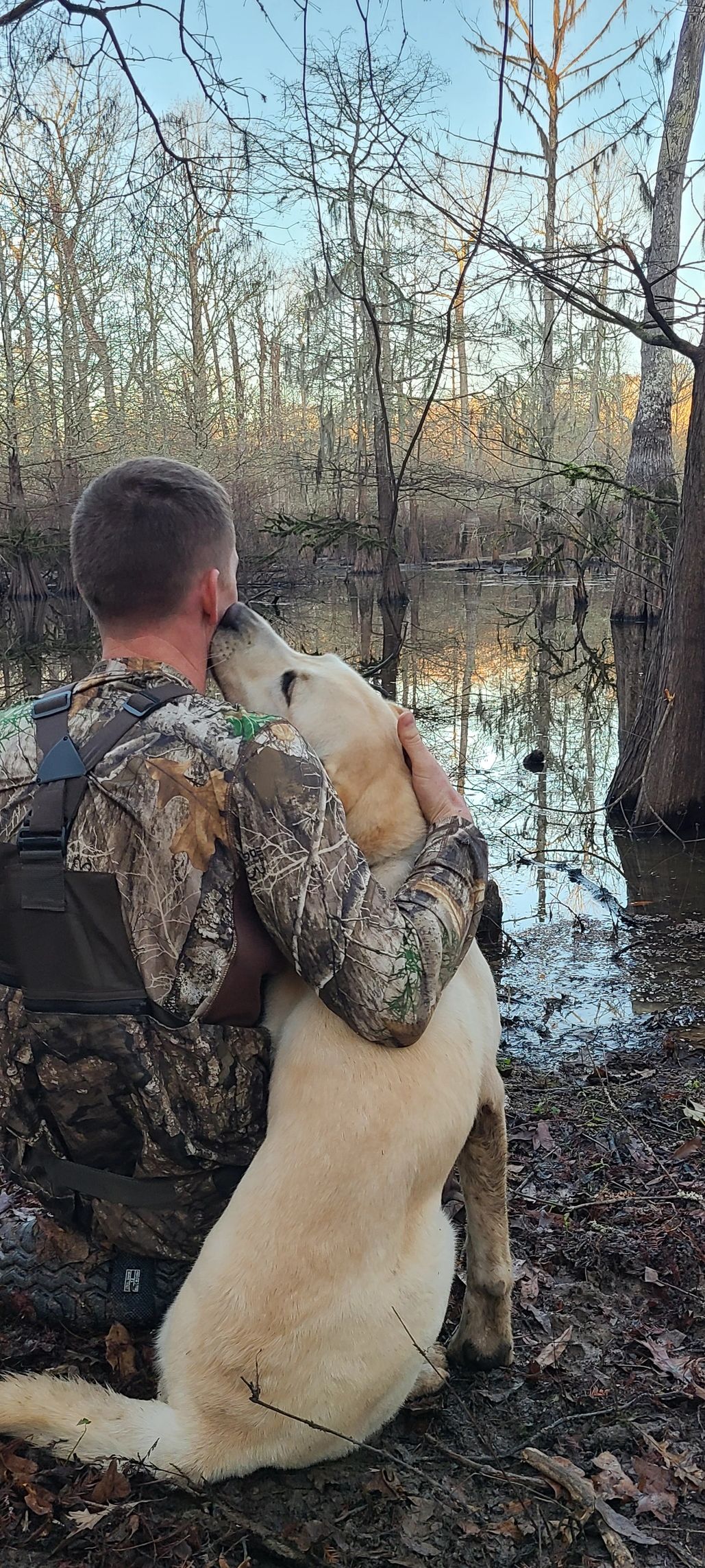 Man in camouflage hugs his dog by a swampy forest.