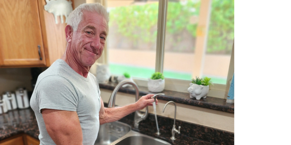 Smiling man in a gray shirt using the kitchen sink faucet.