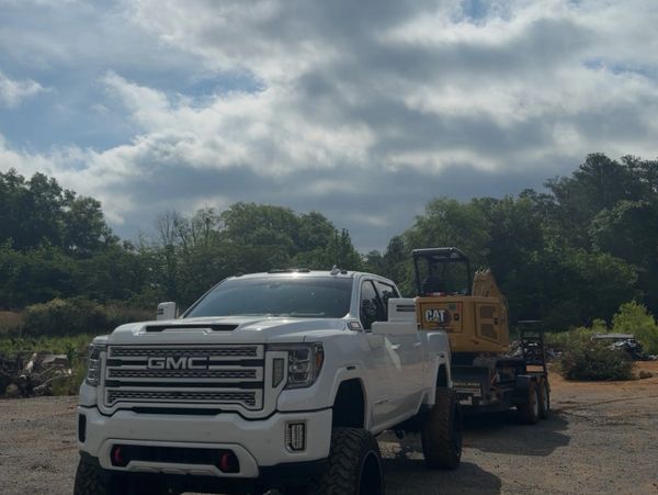 White GMC truck towing a CAT excavator on a trailer in an outdoor setting.