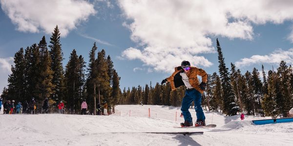 Snowboarder catching air on a snowy slope with trees and spectators in the background.