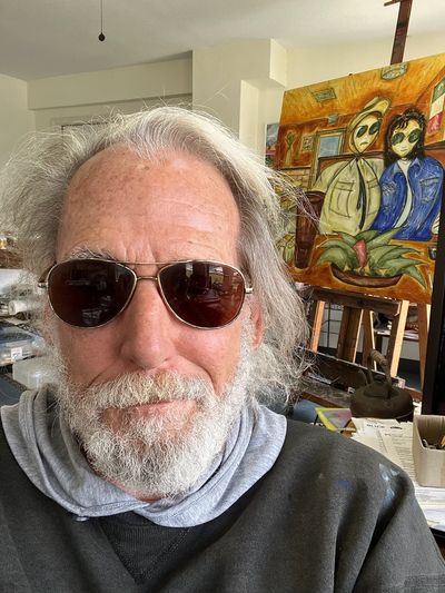 A man with sunglasses and gray hair poses indoors with a colorful painting in the background.