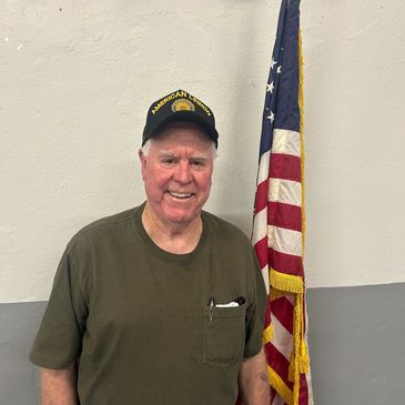 Smiling elderly man in a veteran cap stands beside an American flag.
