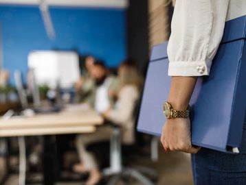 una mujer exhibe en su mano un reloj y sostiene una carpeta azul con documentos