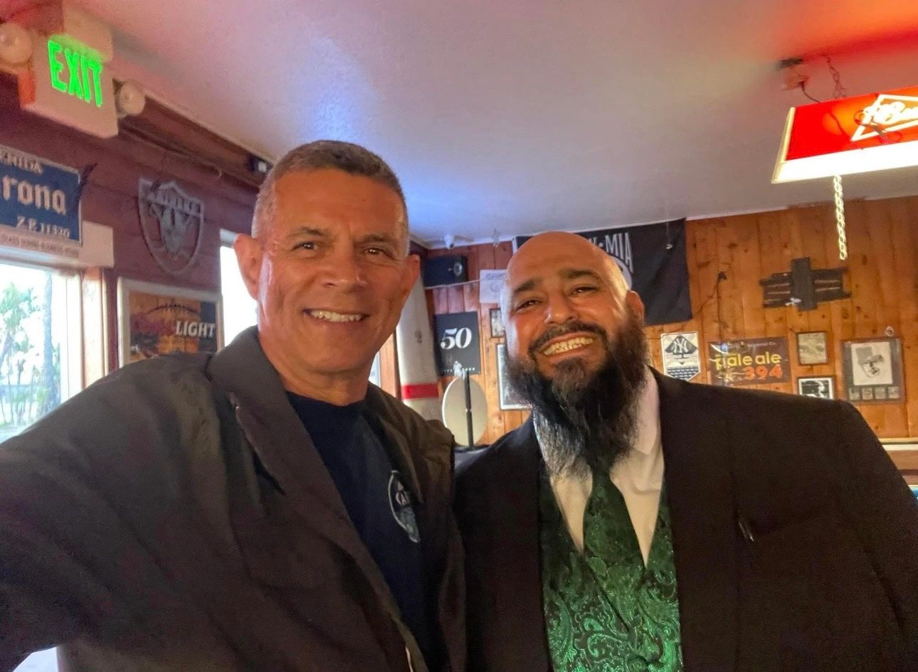 Two men smiling inside a cozy bar with wood-paneled walls.