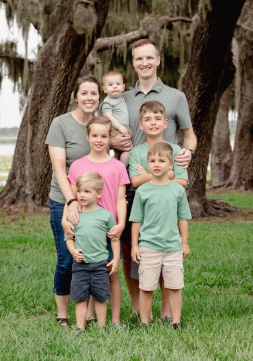 Family of seven posing outdoors under large trees.