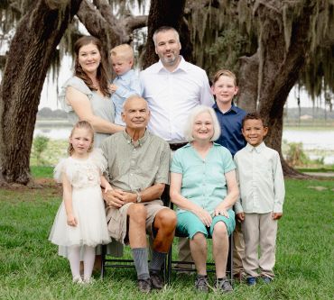 Multi-generational family posing outside under large trees.