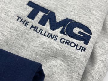 close up of gray shirt with navy blue TMG embroidery