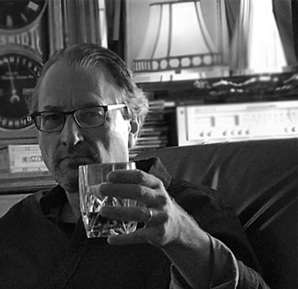 Man with glasses holding a glass, sitting indoors in black and white.