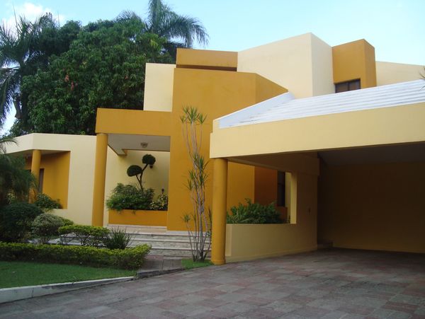 Modern yellow and beige house with a carport and lush greenery.