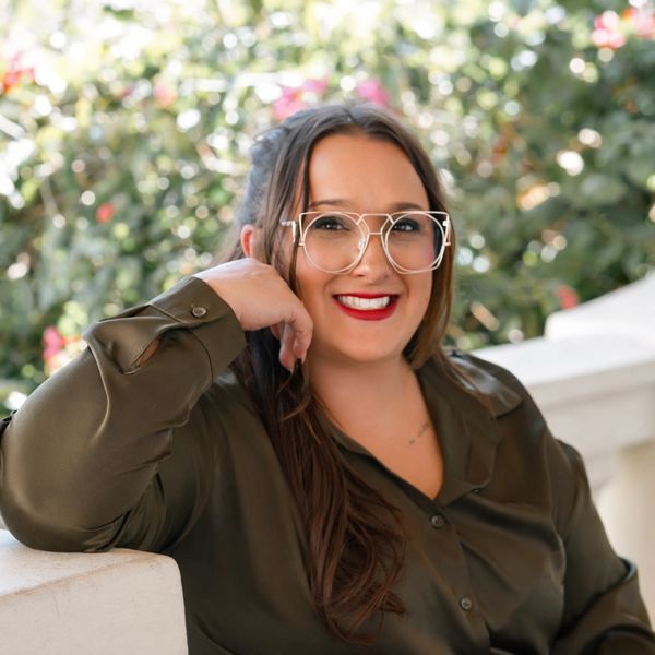 Smiling woman with glasses and red lipstick, leaning on a white railing outdoors.