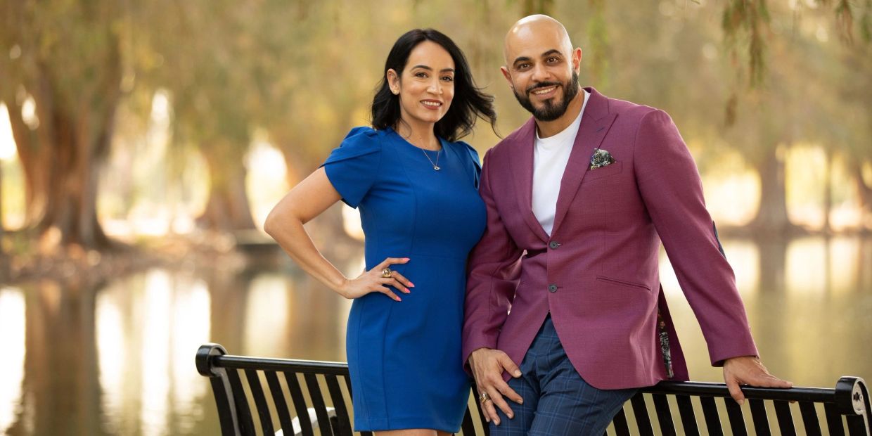 Smiling couple posing by a park bench with serene water and trees in the background.