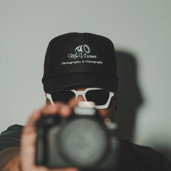 Person wearing a black cap and white sunglasses holding a camera.