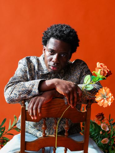 Man leaning on wooden chair holding orange flowers.