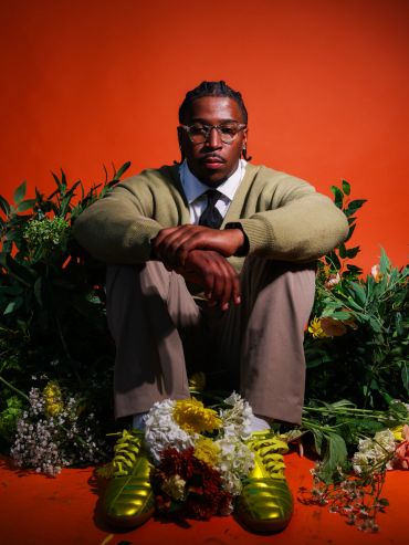 Man with glasses sitting among flowers against an orange background.