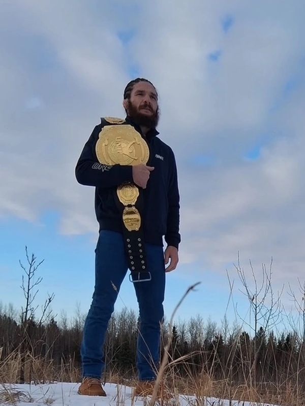 Man proudly holding a championship belt outdoors against a cloudy sky.