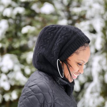 Woman smiling warmly in snowy outdoor setting, wearing a black hood and hoop earrings.