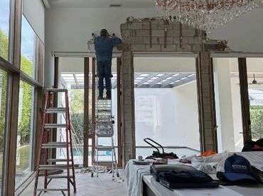 A worker on a ladder repairing a wall inside a bright room.