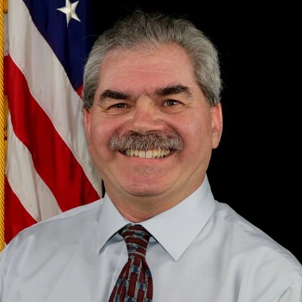 Smiling man in a shirt and tie with an American flag background.