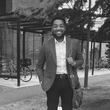 Smiling man in a blazer standing outdoors with a bag.