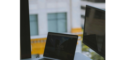 Laptop and monitor setup near a window with city view outside.